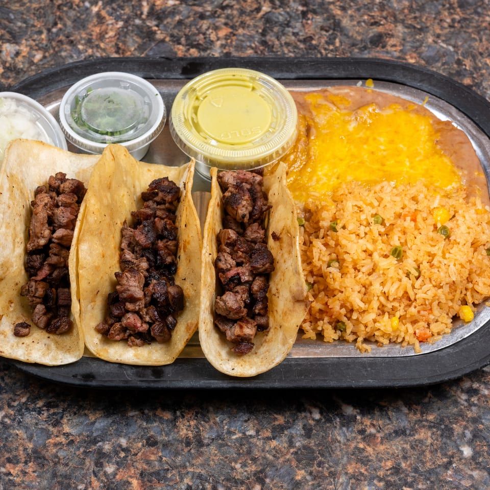 Three grilled meat tacos served with Mexican rice, refried beans, cheese sauce, salsa, and chopped onions.