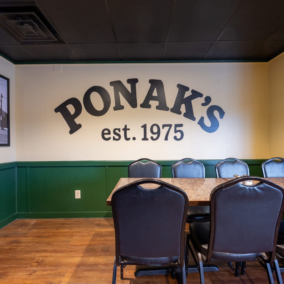 Restaurant dining area with dark green wainscoting, leather chairs, wooden tables, and a wall sign reading “PONAK’S est. 1975.”