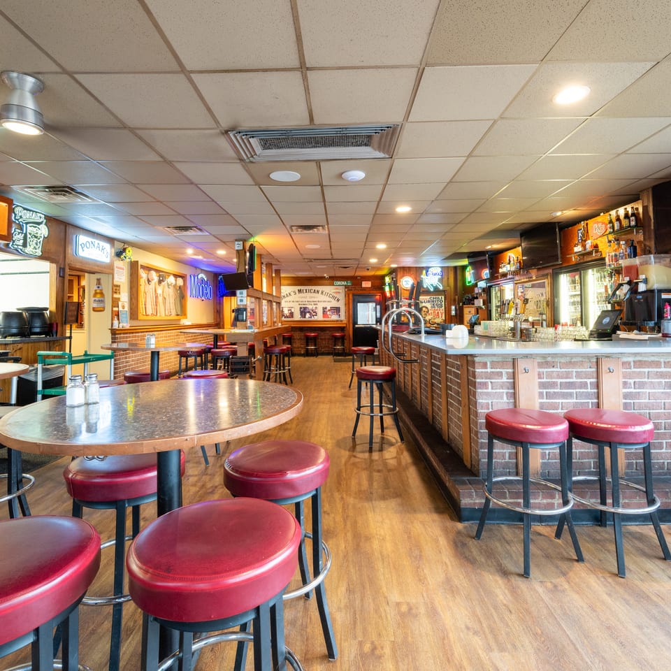 Casual restaurant interior with red bar stools, wooden tables, a brick-front bar, liquor shelves, and colorful wall signs.