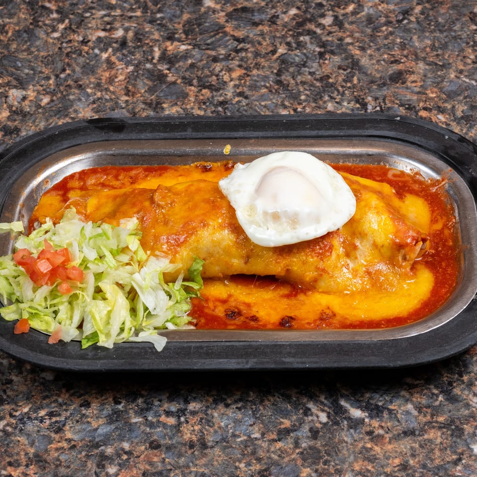 Cheese-covered enchilada topped with a fried egg, served with shredded lettuce and diced tomatoes in a black dish.