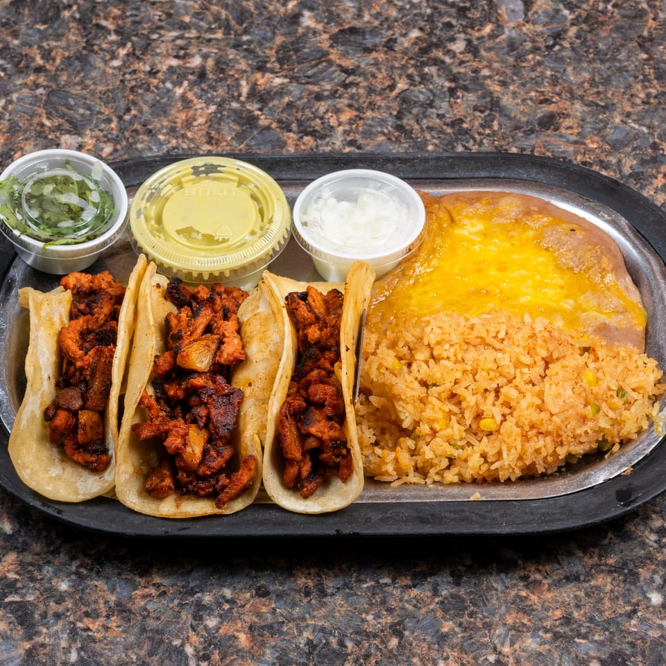 Tray of three tacos filled with grilled meat, Mexican rice, beans, salsa, guacamole, and onions.