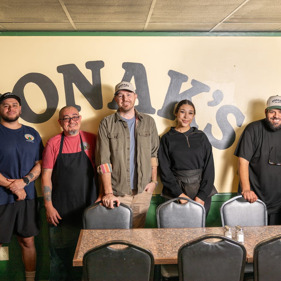 Five restaurant staff members pose together behind a dining table beneath a large painted wall sign.