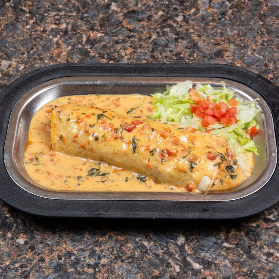 Grilled burrito covered in creamy herb sauce, served with shredded lettuce and diced tomatoes on a black platter.