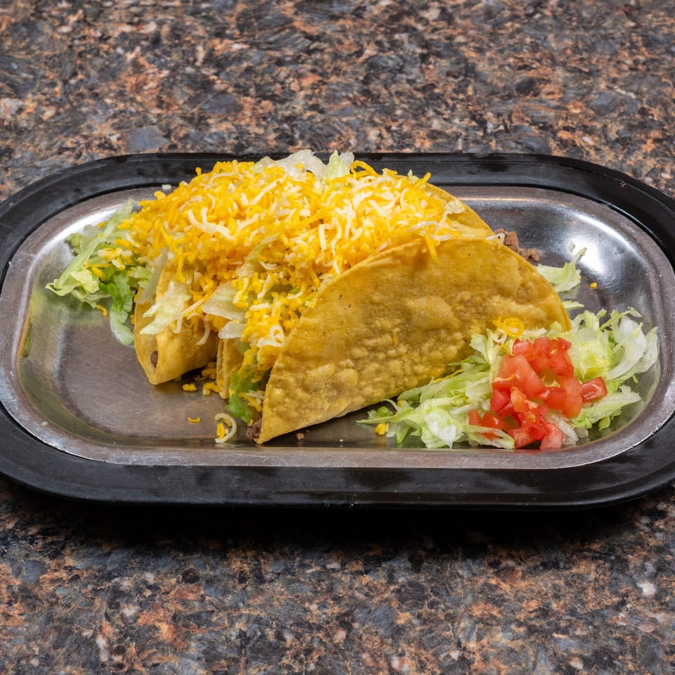 Two crispy beef tacos topped with shredded cheese and lettuce, served with diced tomatoes on a metal tray.