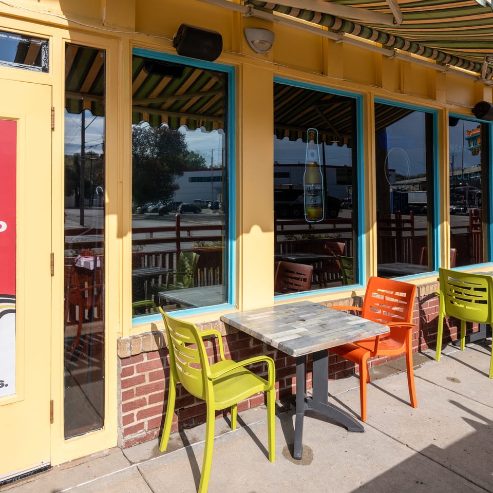 Sunny restaurant exterior with striped awning, turquoise-trimmed windows, outdoor tables, and colorful orange and lime-green chairs.