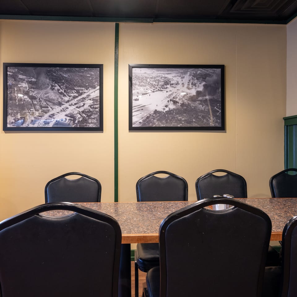 Restaurant dining area with a long table, black chairs, framed aerial photographs, and warm beige walls with green trim.