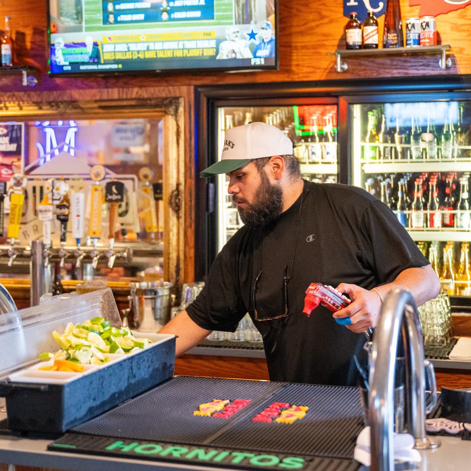 Bartender in a sports bar prepares drinks beside lime wedges, beer taps, and a stocked beverage cooler.