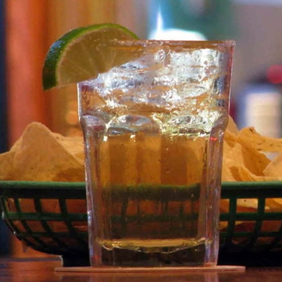 Glass of iced amber beverage garnished with a lime wedge, served beside tortilla chips in a basket.