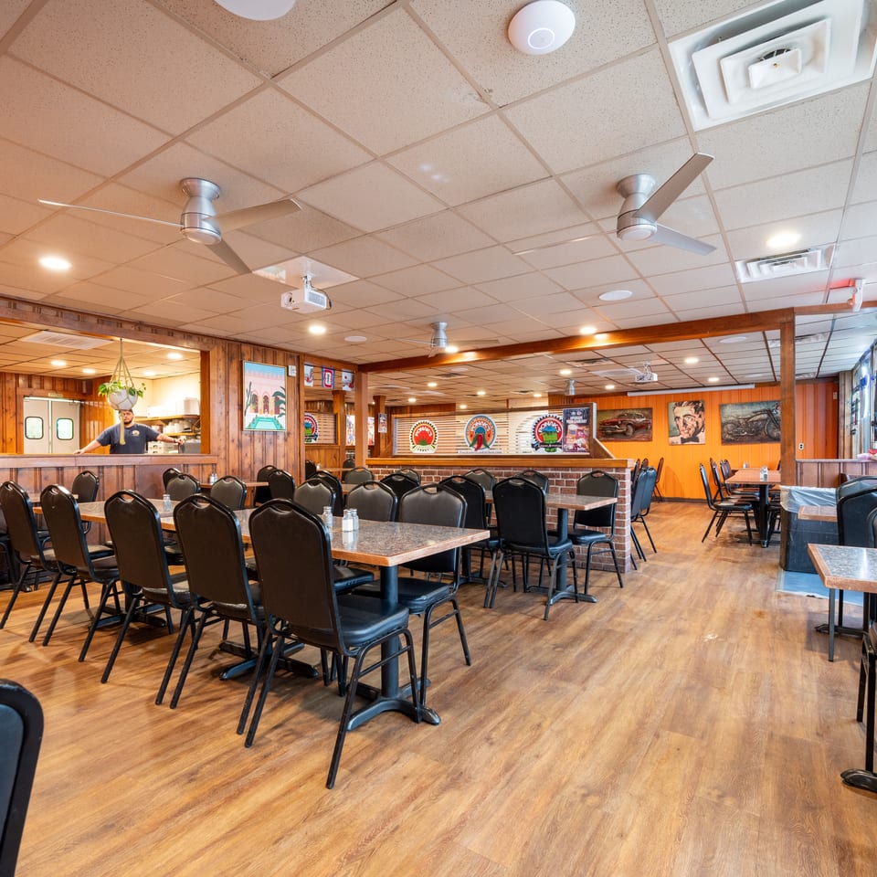 Spacious wood-paneled restaurant dining room with rows of tables, black chairs, ceiling fans, and a server near the counter.