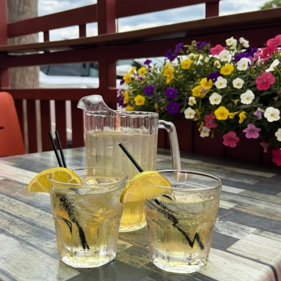 Three glasses of lemon drinks with black straws sit beside a pitcher on a restaurant patio table decorated with colorful flowers.