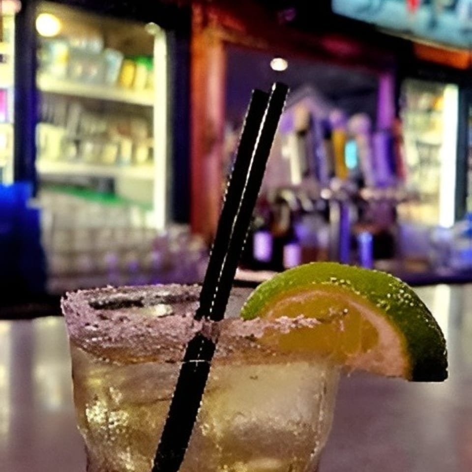 Cocktail with lime garnish and black straw on a coaster at a warmly lit bar.