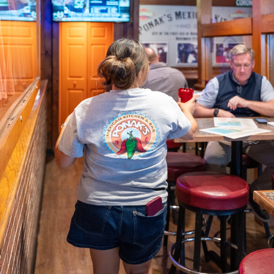 Restaurant interior with a server in a Ponak’s Mexican Kitchen shirt, seated diners, red bar stools, and televisions.