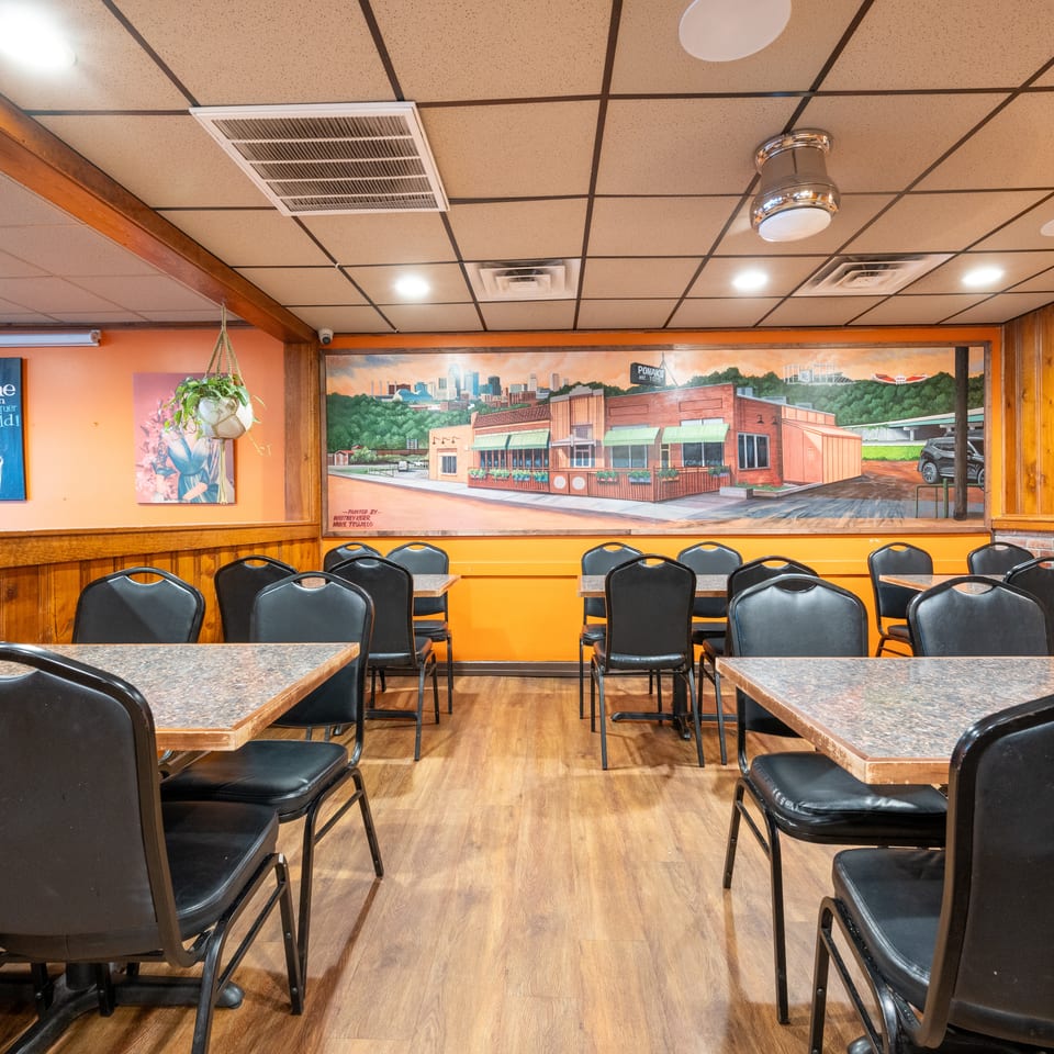 Cozy restaurant dining room with black chairs, wood-paneled walls, colorful artwork, and a large mural of the building exterior.