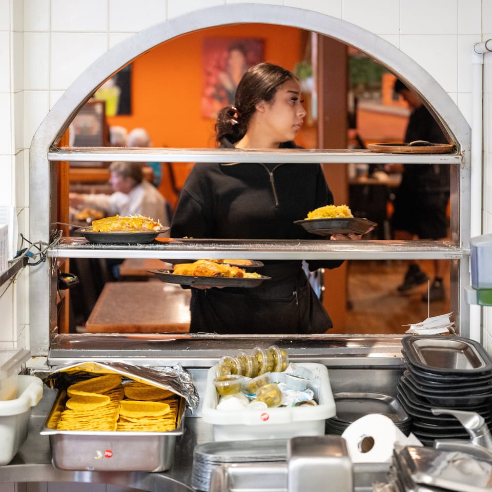 Restaurant worker serves plated dishes through a kitchen pass, with tortillas, toppings, and dining room visible behind her.
